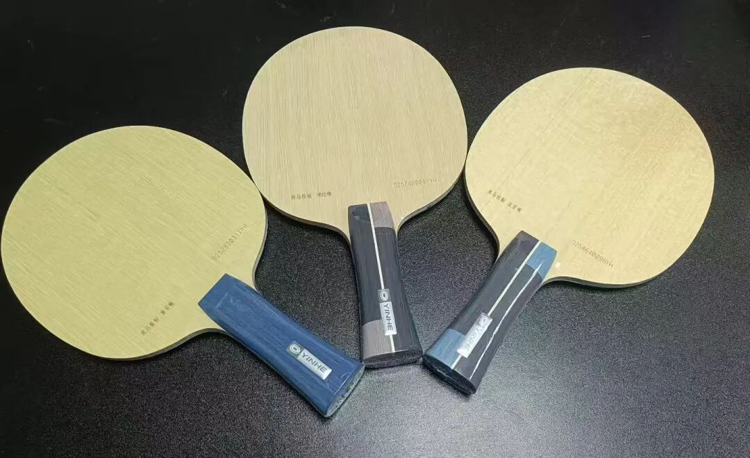 Three table tennis blades laid out on a dark surface showing different handle and face wood grains