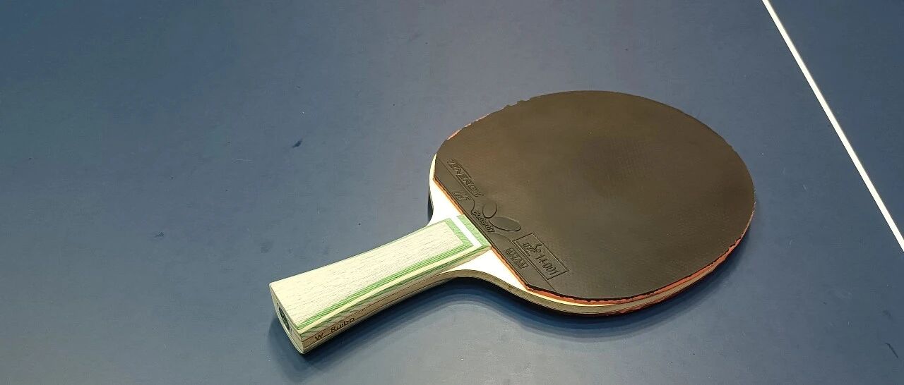 Table tennis paddle on blue table with black rubber, shakehand grip, wood-core blade resting face-down showing handle detail