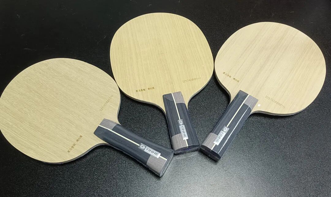 Three Yinhe shakehand blades laid out on a dark surface showing natural wood grain
