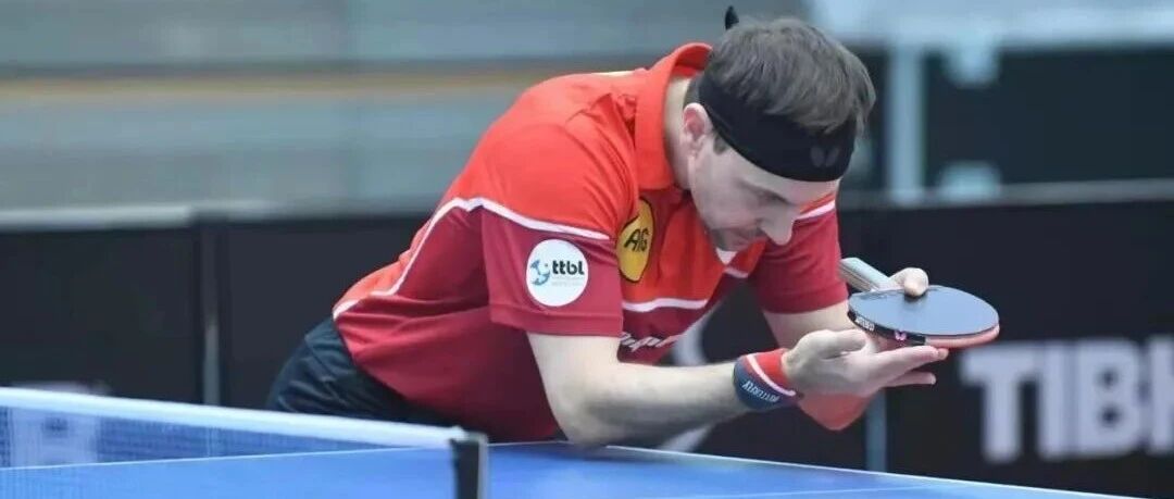 Timo Boll in competition leaning low over the table to serve, wearing red Butterfly jersey with TTBL badge